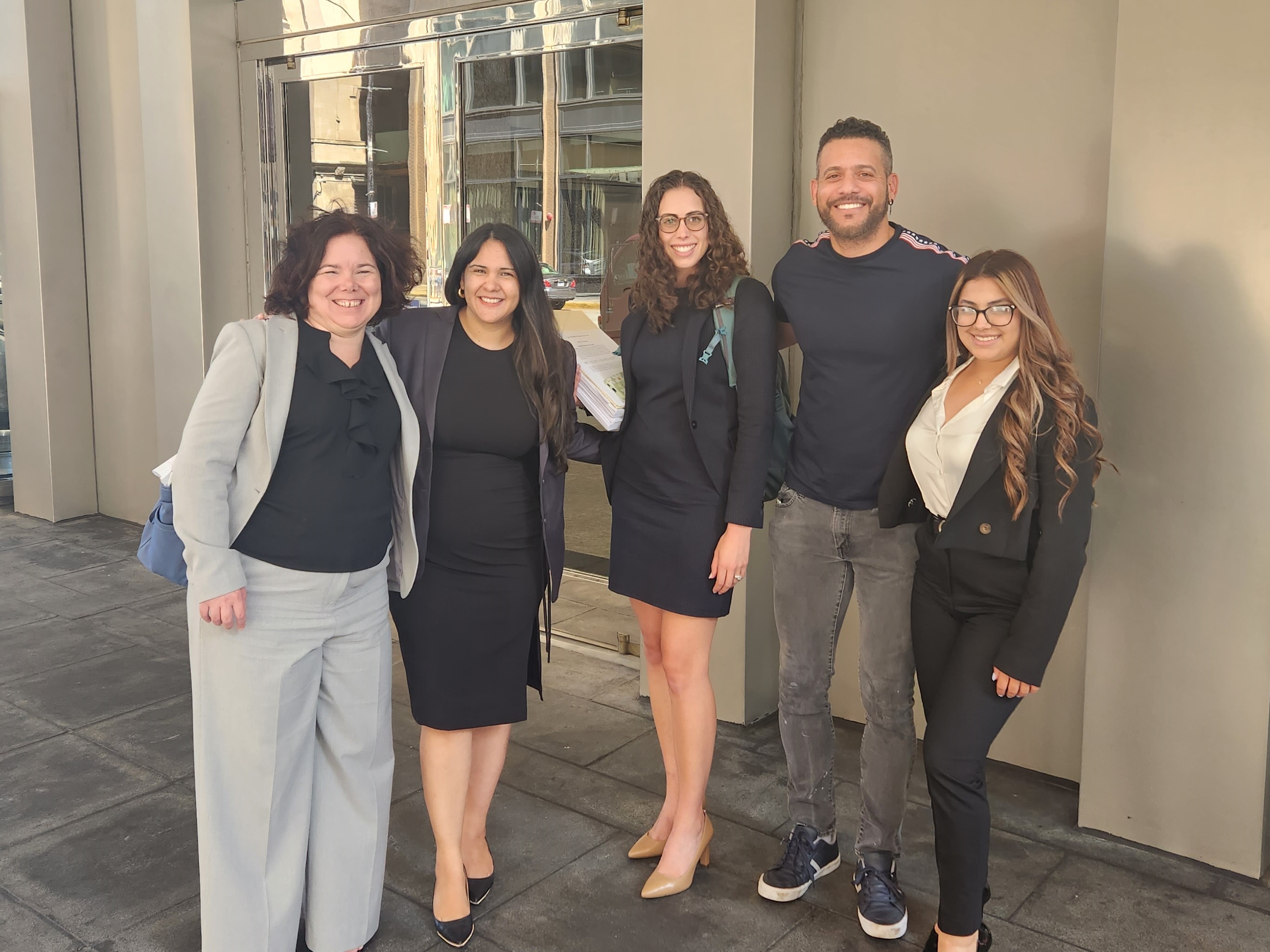 Attorneys and a woman and her friends stand outside of a courthouse, smiling, after the woman won asylum