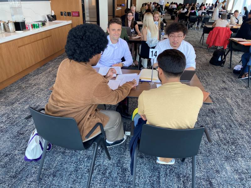 Three young attorneys sit around a table with a client. One of them sits next to the client guiding them through paperwork. They are in a room with lots of other attorneys and clients during a legal clinic.