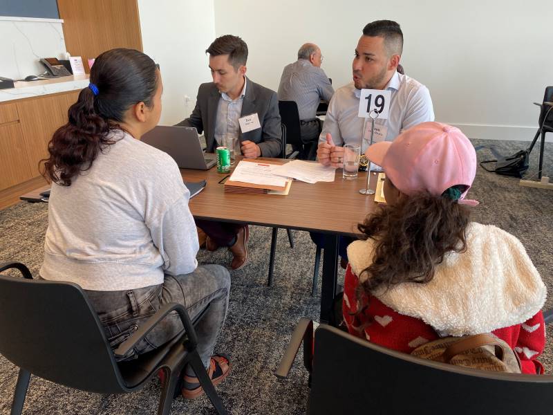 Two attorneys sit across a small table from two women. One attorney is typing on the computer and the other is talking to the women about their legal case.