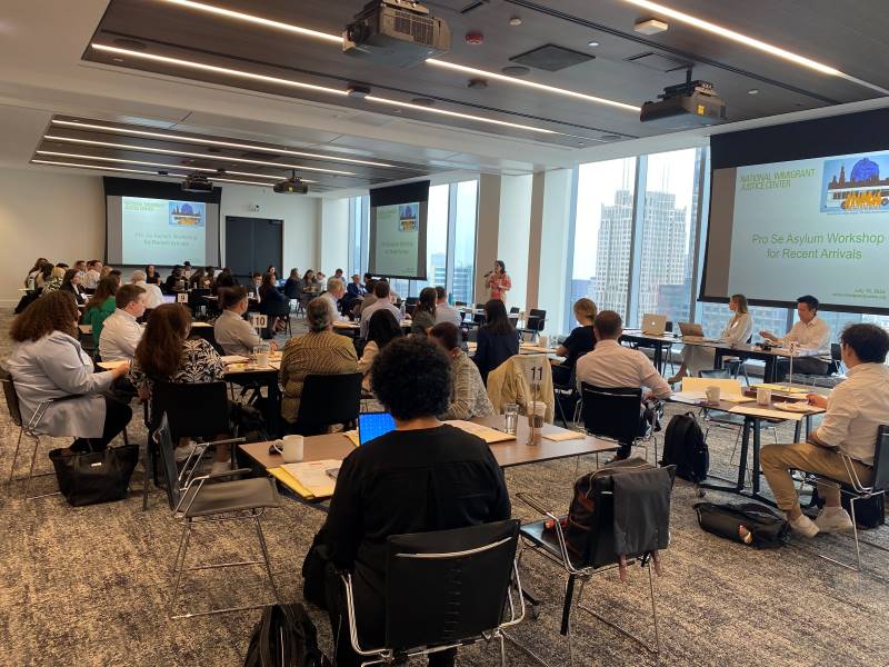 A room full of volunteer attorneys sit in a conference room watching a power point during a training. There are floor to ceiling windows where you can see skyscrapers outside.