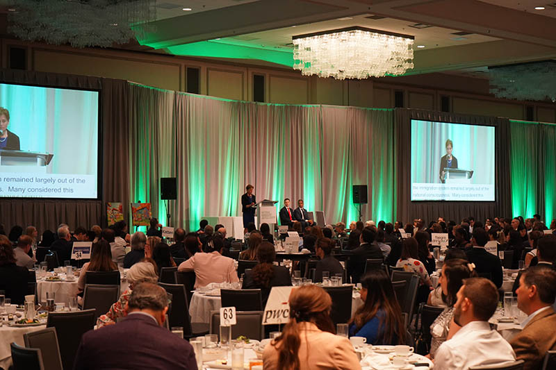 NIJC Executive Director Mary Meg McCarthy speaking at the Human Rights Awards on a stage at the front of a very large ballroom. Many round tables fill the room and event attendees watch her speak. 