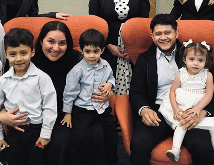 A family sits on a couch smiling, a mother holds her three young sons close and a little girl sits on her father's lap. They are dressed up in nice clothing because they just won asylum in court. Their lawyers stand behind them.