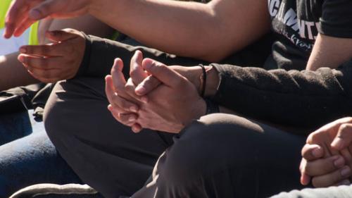 Close-up shot of the torsos and clasped hands of three people sitting next on the ground next to each other, their legs crossed.