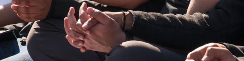 Close-up shot of the torsos and clasped hands of three people sitting next on the ground next to each other, their legs crossed.