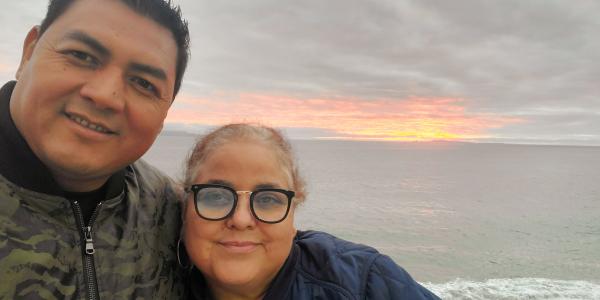 Photo of Luis Asencio Cordero and Sandra Munoz, whose case regarding due process rights for spouses in mixed-status marriages is before the U.S. Supreme Court. Luis has his arm around Sandra and both are smiling at the camera. Behind them the sun is setting over a body of water.